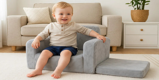 A smiling toddler sits on a gray 2-in-1 plush kids’ sofa chair with the footrest extended in a bright, cozy living room.