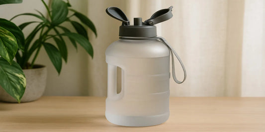 A frosted half-gallon water bottle with a gray flip-top lid and fabric strap, placed on a wooden table with plants and curtains in the background.