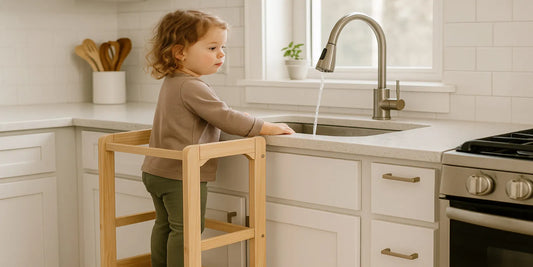 A toddler stands on a wooden kitchen step stool at the sink, reaching toward running water in a bright modern kitchen with white cabinets.