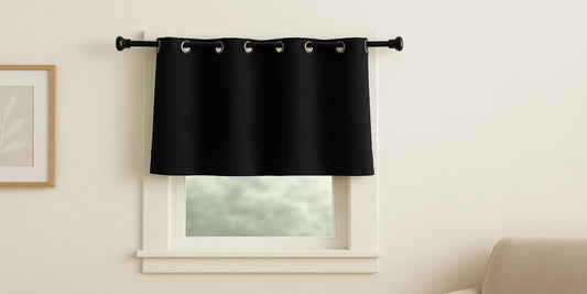 A short black blackout curtain hanging on a small window, blocking light while allowing a soft view of greenery outside the room.